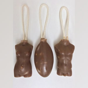 May include: Three brown soap bars in the shape of a female torso, an oval, and a male torso, each with a white rope loop for hanging. The soaps are arranged against a white background.