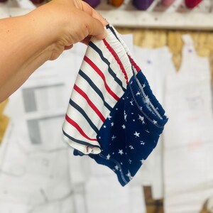 May include: A pair of baby shorts with a patriotic design. One side features red and white stripes, while the other has a navy blue background with white stars. The shorts are held up by a hand, with spools of thread in the background.