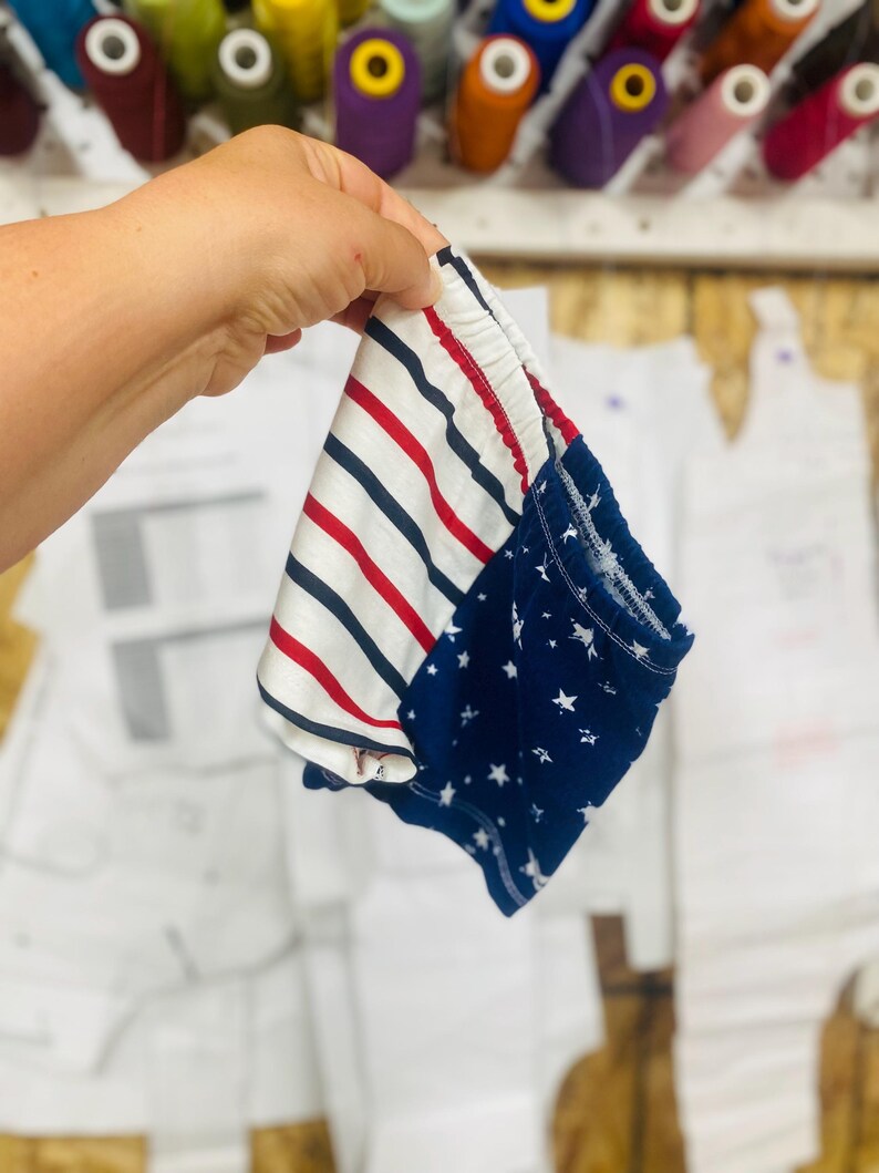 May include: A pair of baby shorts with a patriotic design. One side features red and white stripes, while the other has a navy blue background with white stars. The shorts are held up by a hand, with spools of thread in the background.