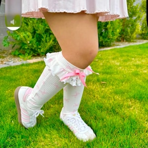 May include: White knee-high socks with a pink floral pattern and ruffled lace trim. The socks have a pink bow detail and are paired with white sneakers with pink stripes and brown soles. The image is set against a backdrop of green grass.
