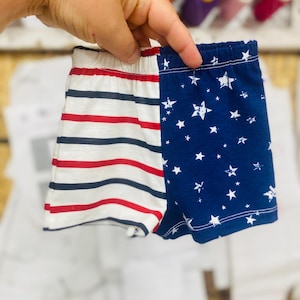 May include: A pair of baby shorts with a patriotic design. One side features red and navy blue stripes on a white background, while the other side has a navy blue background with white stars. The shorts have an elastic waistband.