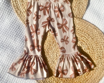 May include: Cream-colored baby pants with a brown football and bow pattern. The pants have a ribbed texture and flared bottoms. The design features repeating football and bow illustrations.