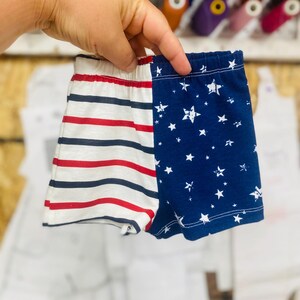 May include: A pair of baby shorts, one side with red and navy stripes, the other with a navy blue background and white stars. The shorts are held up by a hand, with spools of thread in the background.