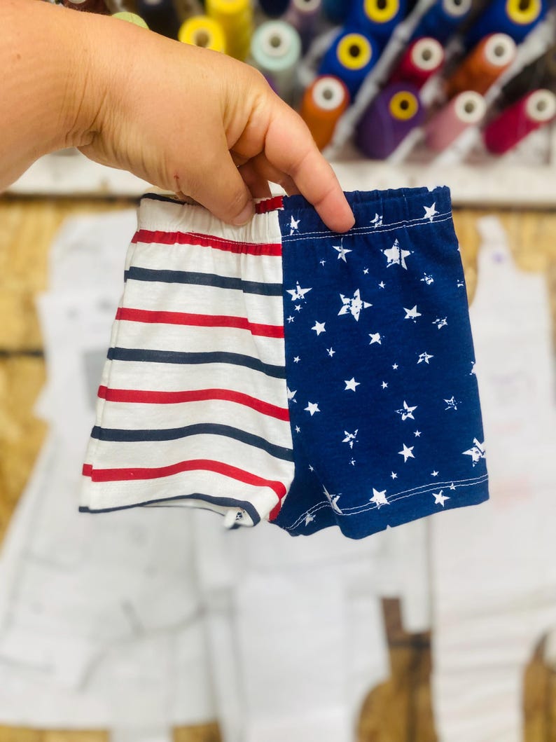May include: A pair of baby shorts, one side with red and navy stripes, the other with a navy blue background and white stars. The shorts are held up by a hand, with spools of thread in the background.