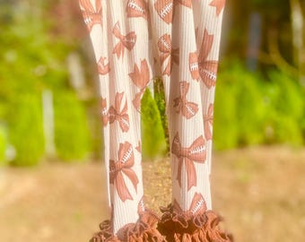 May include: Cream-colored leggings with a repeating pattern of brown football-shaped bows. The leggings have a ribbed texture and are finished with ruffled trim at the ankles. The background is blurred, suggesting an outdoor setting.