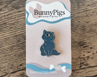 May include: A blue cat enamel pin with a silver outline, sitting on a light pink and blue card. The card has the text "BunnyPigs enamel pins" and "bunnypigs.com". The pin is on a wooden surface.