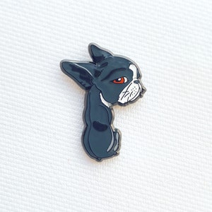 May include: A black and white Boston Terrier pin with red eyes. The pin is a cartoon-style illustration of the dog in a sitting position, with its head turned to the side. The pin has a glossy finish.