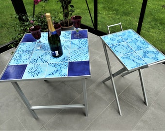 May include: Two blue and white tiled tables with silver metal frames. The larger table has a bottle of champagne and two wine glasses on it. The smaller table is a folding table.