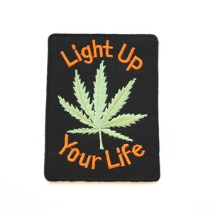 Puede incluir: Parche bordado negro con una hoja de marihuana verde y el texto "Light Up Your Life" en naranja.