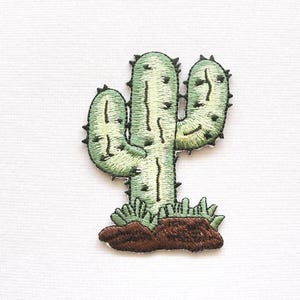 Könnte beinhalten: Gestickter Kaktus-Aufnäher in Grün, Braun und Schwarz. Der Kaktus hat ein detailliertes Design mit Stacheln und ist vor einem weißen Hintergrund platziert. Der Aufnäher ist für Kleidung oder Accessoires geeignet.