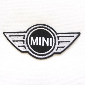 Op de afbeelding: Geborduurde patch met het MINI-logo. De patch heeft een zwart cirkelvormig midden met het woord "MINI" in wit, omlijnd door een witte rand en geflankeerd door gestileerde vleugels. De vleugels en tekst zijn wit op een zwarte achtergrond.