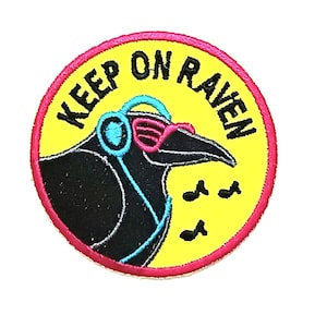 Peut inclure: Un patch rond jaune et rose avec un corbeau noir portant un casque et des lunettes de soleil. Le texte "KEEP ON RAVEN" est écrit autour du corbeau.