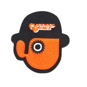 Puede incluir: Parche bordado en negro y naranja con un personaje de la película "La naranja mecánica". El personaje lleva un sombrero bombín y tiene un ojo grande y redondo con un engranaje.