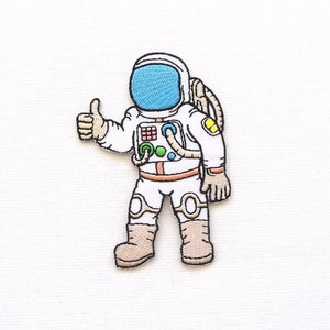 Puede incluir: Un parche bordado para planchar de un astronauta de dibujos animados que lleva un traje espacial blanco con una visera azul, haciendo un pulgar hacia arriba.