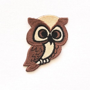 Könnte beinhalten: Brauner gestickter Eulen-Patch mit schwarzen Augen und einem beigefarbenen Kopf. Die Eule blickt nach links und hat ihre Flügel ausgebreitet.