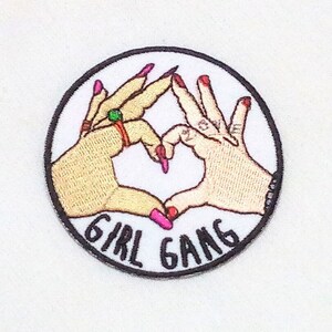 Può includere: Toppa ricamata da stirare con due mani che formano un cuore. Le mani hanno lo smalto rosa e il testo "Girl Gang" è scritto in nero sotto il cuore.
