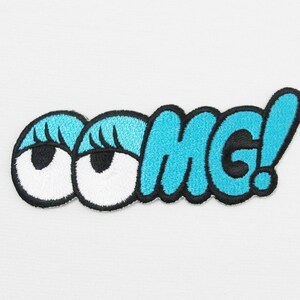 Puede incluir: Un parche bordado azul y negro con la palabra "OMG!" en una fuente de dibujos animados. Dos grandes ojos de dibujos animados con pestañas están en el lado izquierdo del parche.