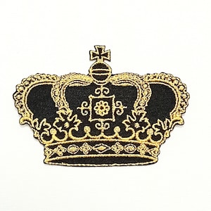 Può includere: Un toppa ricamata a forma di corona nera e dorata. La corona ha una cima appuntita con una croce e dettagli intricati.
