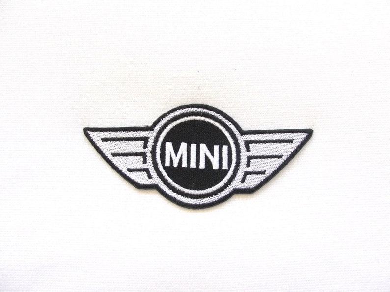 Pode incluir: Patch bordado com o logotipo MINI. O patch tem um centro circular preto com a palavra "MINI" em branco, emoldurado por um contorno branco e ladeado por asas estilizadas. As asas e o texto s&atilde;o brancos sobre um fundo preto.
