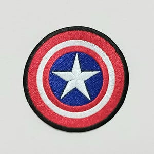 May include: Embroidered patch featuring a circular design with a red outer ring, white stripes, a blue center, and a white star. The patch has a black border and is likely for clothing or accessories.