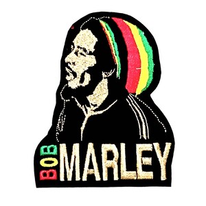 Puede incluir: Parche bordado en negro y dorado con un perfil de Bob Marley con un sombrero a rayas rojo, amarillo y verde. El texto "BOB MARLEY" está bordado en dorado debajo de la imagen.