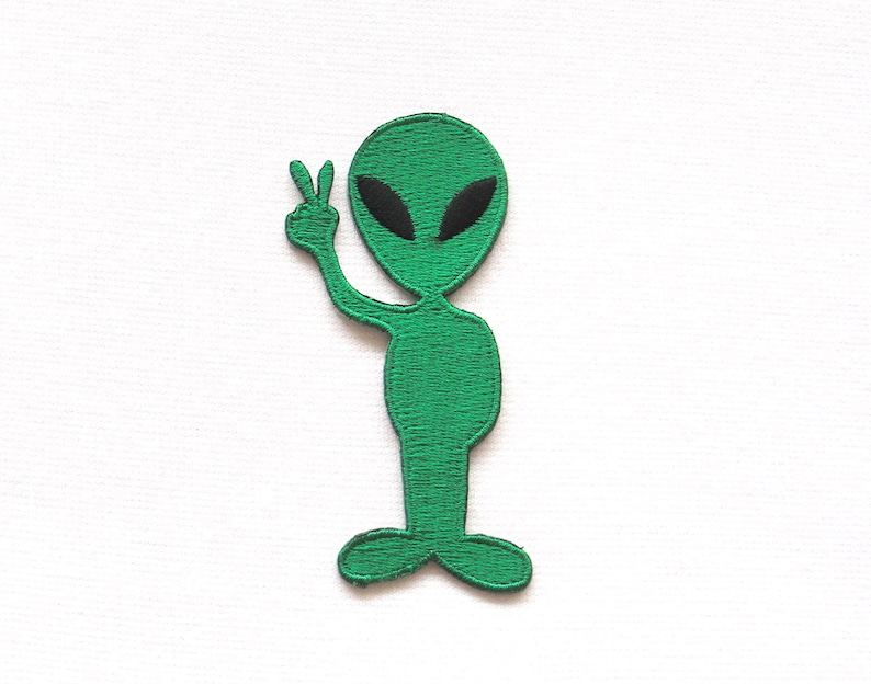 May include: Green alien with black eyes holding up two fingers in a peace sign. The alien is standing on a white background.