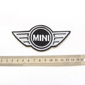 Pode incluir: Um patch bordado em preto e branco com a palavra "MINI" dentro de um emblema circular com extens&otilde;es em forma de asa. Uma r&eacute;gua de metal com marca&ccedil;&otilde;es em cent&iacute;metros est&aacute; colocada abaixo.