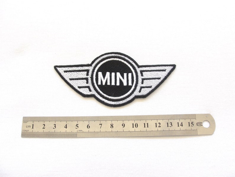 Pode incluir: Um patch bordado em preto e branco com a palavra "MINI" dentro de um emblema circular com extens&otilde;es em forma de asa. Uma r&eacute;gua de metal com marca&ccedil;&otilde;es em cent&iacute;metros est&aacute; colocada abaixo.