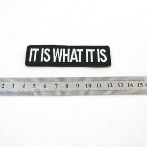 May include: Black and white embroidered patch with the text "IT IS WHAT IT IS" on a black background. The patch is approximately 6 cm long.