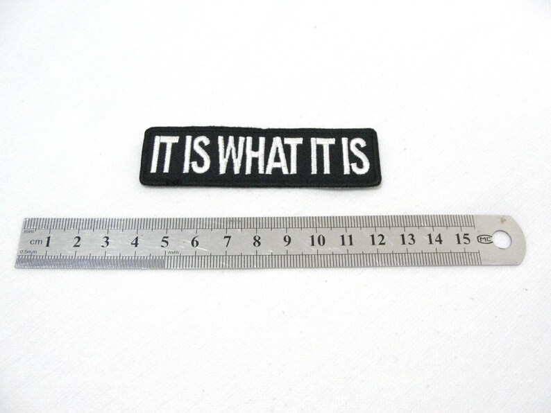 May include: Black and white embroidered patch with the text "IT IS WHAT IT IS" on a black background. The patch is approximately 6 cm long.