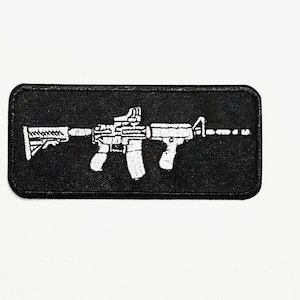 Könnte beinhalten: Schwarzer rechteckiger Aufnäher mit einem weißen, gestickten Gewehrdesign. Das Gewehr ist detailliert und stilisiert dargestellt, mit sichtbaren Komponenten. Der Aufnäher hat ein strukturiertes Aussehen. Militärisches Thema.