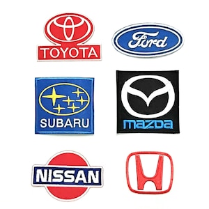May include: A collection of six embroidered patches featuring automotive brand logos. The set includes the red Toyota logo, blue Ford oval, blue Subaru emblem, black Mazda badge, red and blue Nissan logo, and the red Honda H-mark emblem.