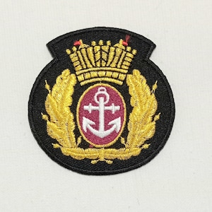 Könnte beinhalten: Gestickter Aufnäher mit goldener Krone, Lorbeerblättern und Ankeremblem auf schwarzem Hintergrund. Der Anker ist weiß, in einem bordeauxfarbenen Kreis. Der Aufnäher hat eine schildförmige Form.