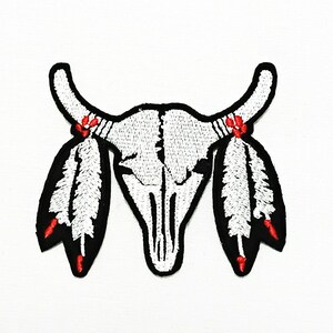 May include: Embroidered patch featuring a white bull skull with black outlines. Two white and black feathers with red accents are attached to the horns. The patch is designed with detailed stitching, suitable for application on clothing or accessories.
