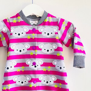 May include: A pink and gray long-sleeved baby onesie with a koala bear print and yellow stars. The onesie has a button closure down the front.