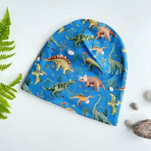 Peut inclure: Un bonnet bleu à motif de dinosaures. Le bonnet présente une variété de dinosaures de différentes couleurs et poses, notamment un stégosaure, un tricératops et un ptérodactyle.