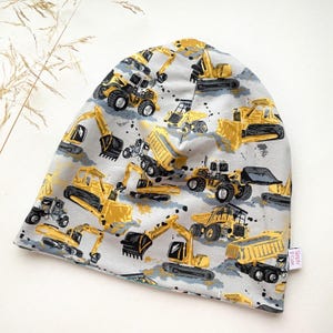 May include: Grey beanie with a yellow and black construction vehicle print. The beanie has a fold-over cuff and the text "Simply & Cute" is printed on a white tag.