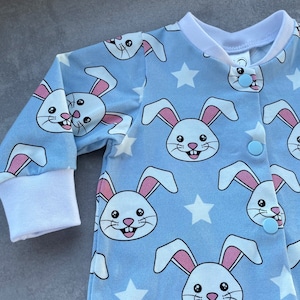 Puede incluir: Un pelele de bebé azul claro con un estampado de conejito blanco y botones azules. El pelele tiene mangas largas y un cuello blanco.