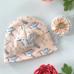 May include: Peach-colored beanie hat with a white hummingbird pattern and white squares. The hat has a tag that says "Simply 2 Cute" and a button that says "Handmade with love".