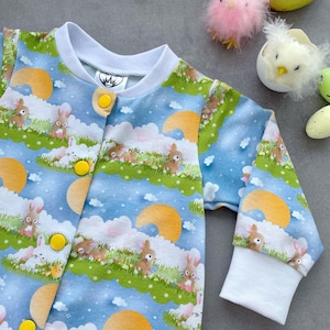 Puede incluir: Mono de bebé con estampado azul y blanco con un diseño caprichoso de conejito de Pascua y paisaje. La prenda de manga larga tiene cierres de botón amarillos y puños y cuello blancos. El diseño incluye conejitos, nubes y soles.