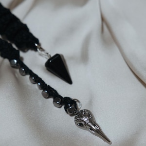 May include: Close-up of a black and silver pendant necklace. The necklace features a black, cone-shaped pendant and a silver bird skull pendant. The cord is black and has small, silver beads.