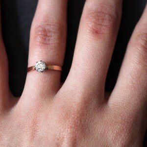 May include: A close-up of a hand wearing a gold ring with a single, round diamond.