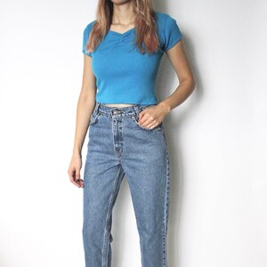 May include: A woman wearing a light blue cropped t-shirt and blue denim jeans. She is wearing brown sandals.
