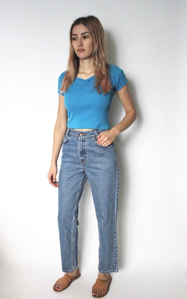 May include: A woman wearing a light blue cropped t-shirt and blue denim jeans. She is wearing brown sandals.
