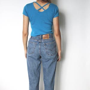 May include: A person wearing a blue short-sleeved t-shirt with a criss-cross back detail and blue denim jeans.