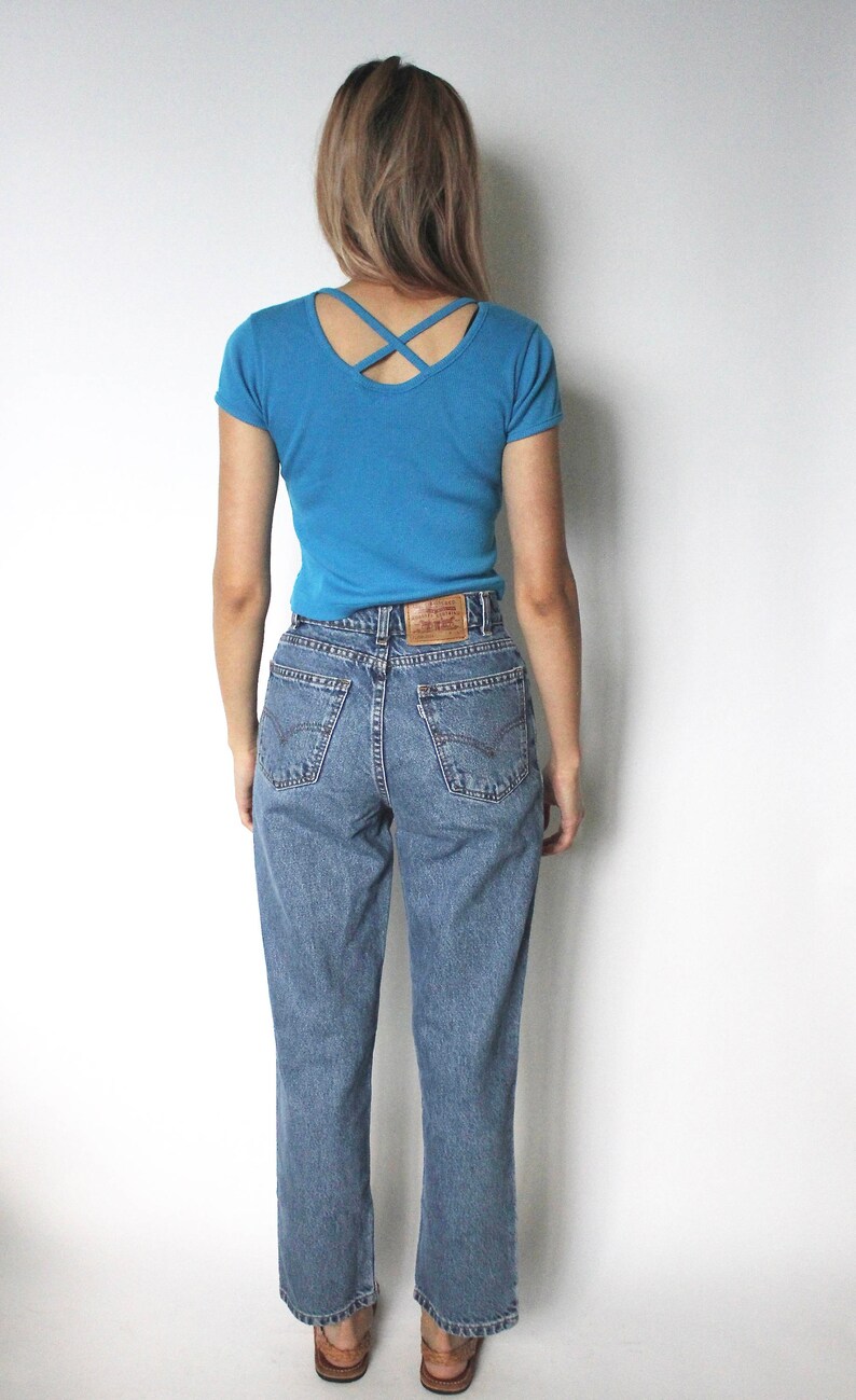 May include: A person wearing a blue short-sleeved t-shirt with a criss-cross back detail and blue denim jeans.