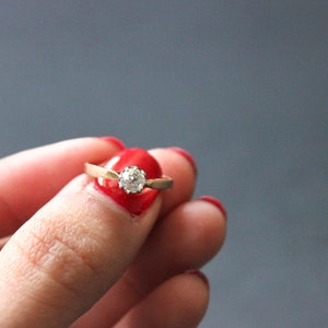 May include: A gold ring with a single round diamond set in the center. The ring is on a finger with red nail polish.