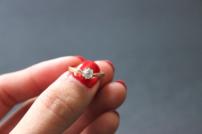 May include: A gold ring with a single round diamond set in the center. The ring is on a finger with red nail polish.