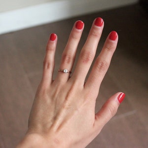 May include: A hand with red nail polish is wearing a gold ring with a single diamond. The hand is against a light brown background.