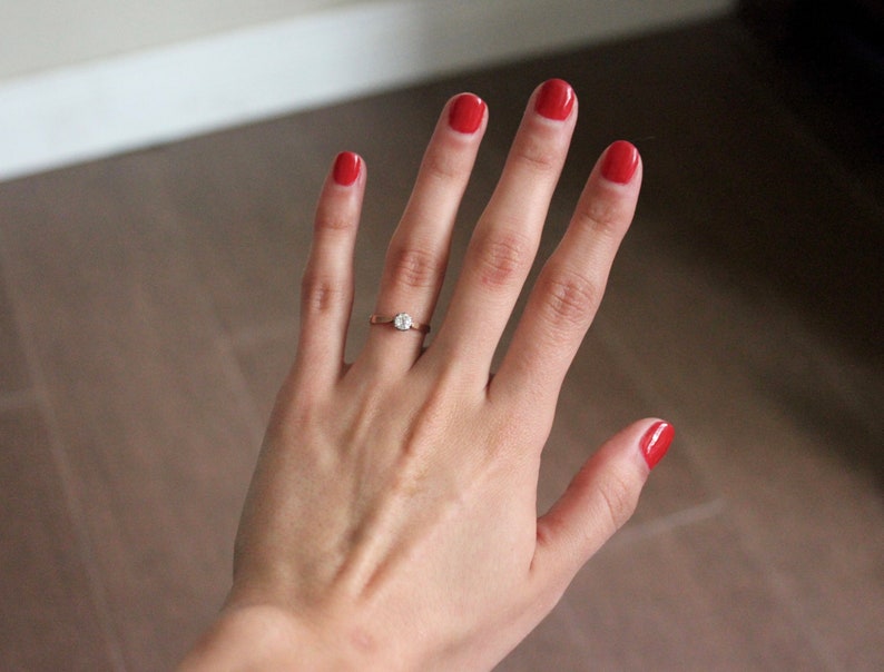 May include: A hand with red nail polish is wearing a gold ring with a single diamond. The hand is against a light brown background.